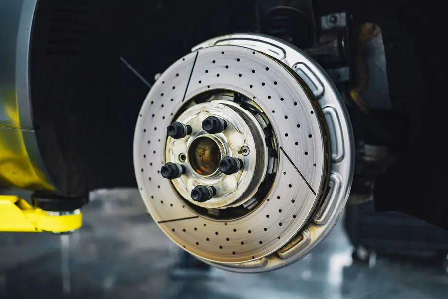 A drilled brake rotor with its wheel removed, mounted on a car raised on a lift in a dim workshop.