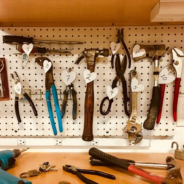 Hand tools including pliers, a hammer and wrenches hanging on a white pegboard above a wooden workbench.